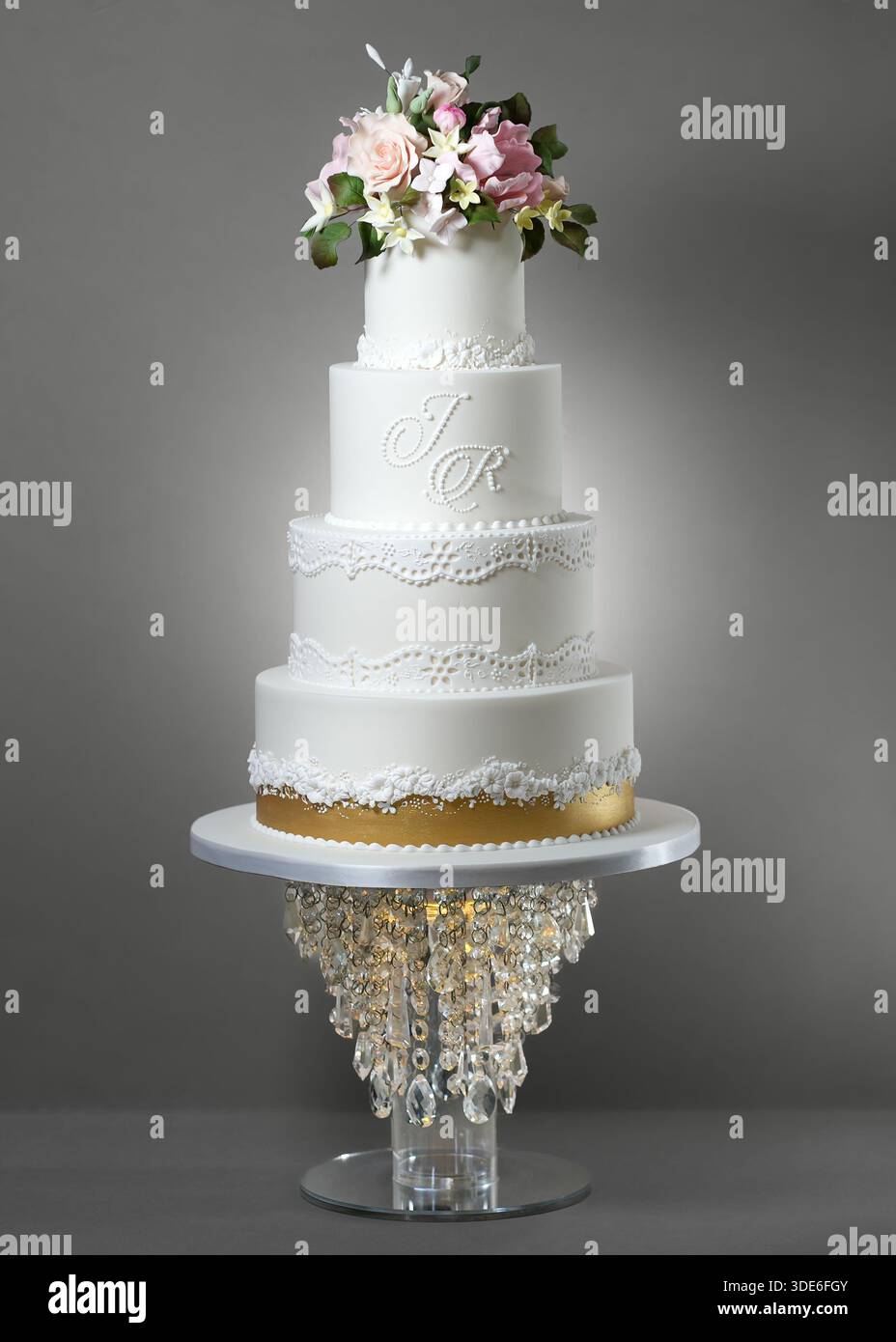 Elegante, vierstöckige weiße Hochzeitstorte mit Spitzenpaspeln, Blumendetails, goldenem Akzentband und Monogramm mit pastellfarbenem Zuckerfleck Stockfoto