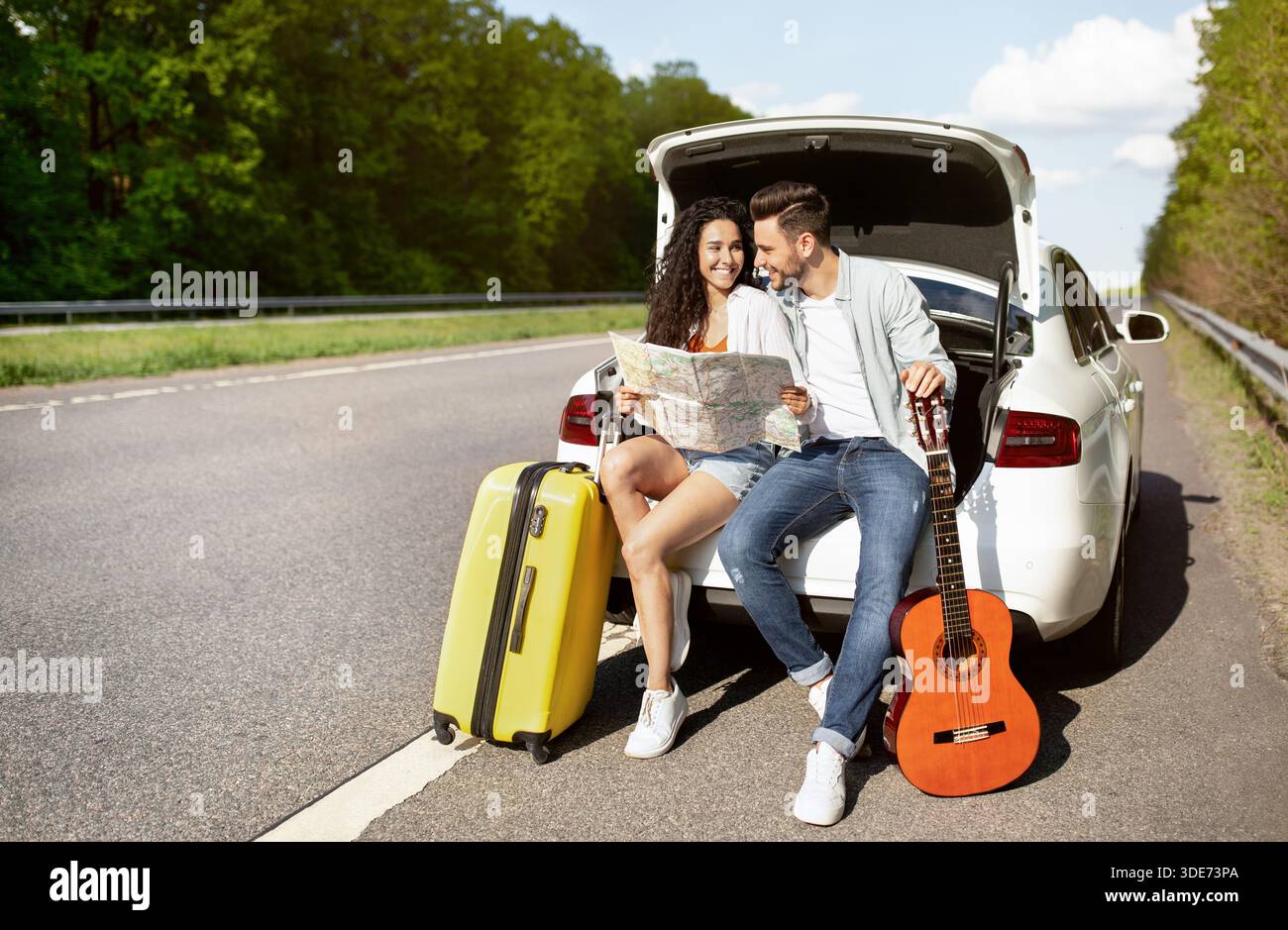 Junges Paar, das mit dem Auto unterwegs ist, im Kofferraum mit Koffer und Gitarre sitzt, Karte hält, Ziel überprüft Stockfoto