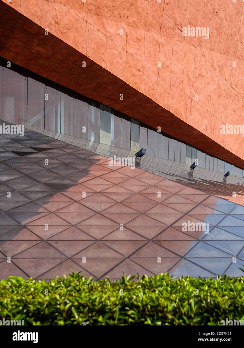Abstrakte Ansicht einer Gebäudeecke mit strukturierter roter Wand, grauem, geometrischem Pflaster und grüner Hecke. Das Bild konzentriert sich auf Linien Stockfoto