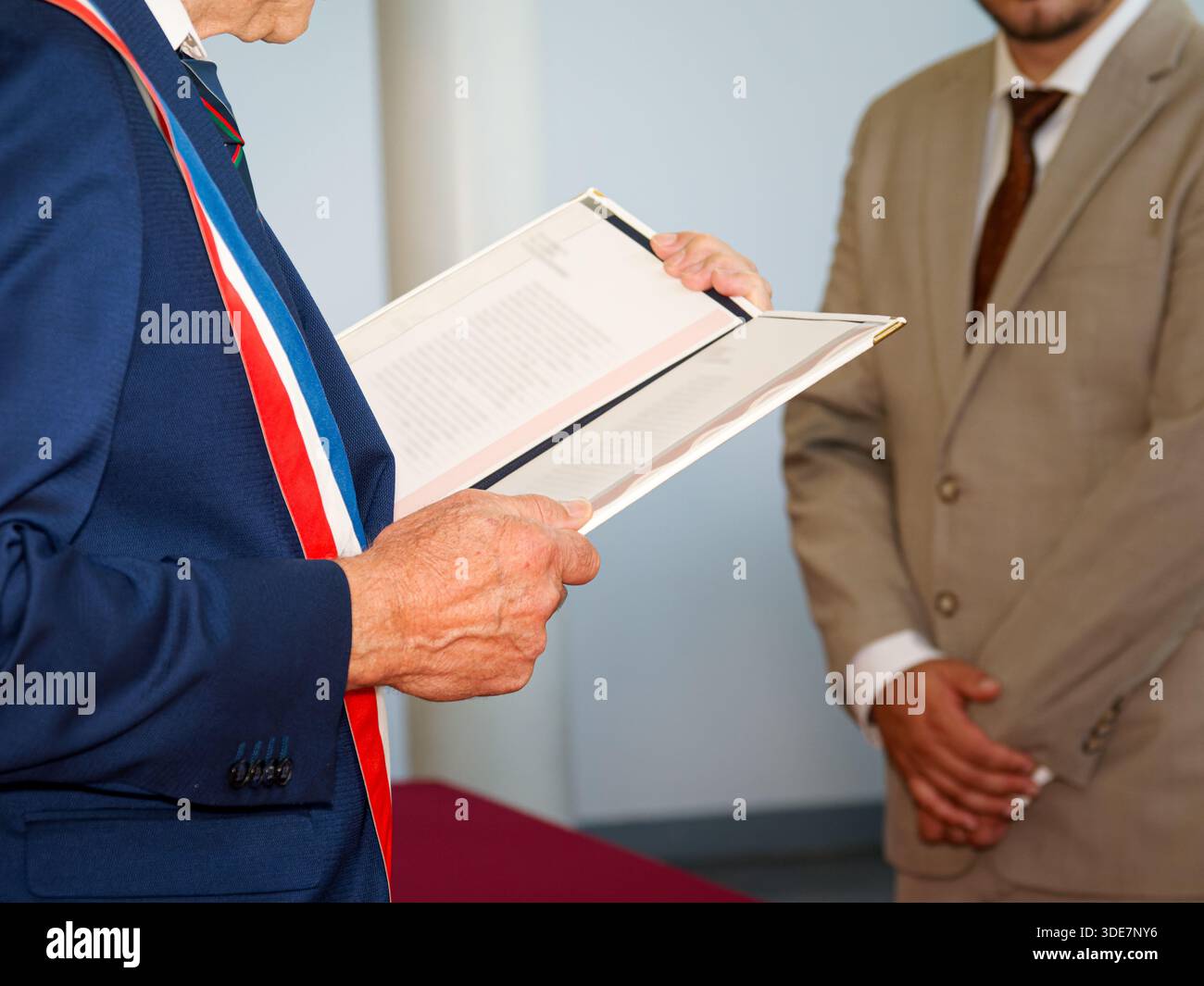Der französische Bürgermeister mit einem dreifarbigen Schärpe liest während einer Bürgerzeremonie aus einem offiziellen Register, während ein anderer Beamter in der Nähe steht. Stockfoto