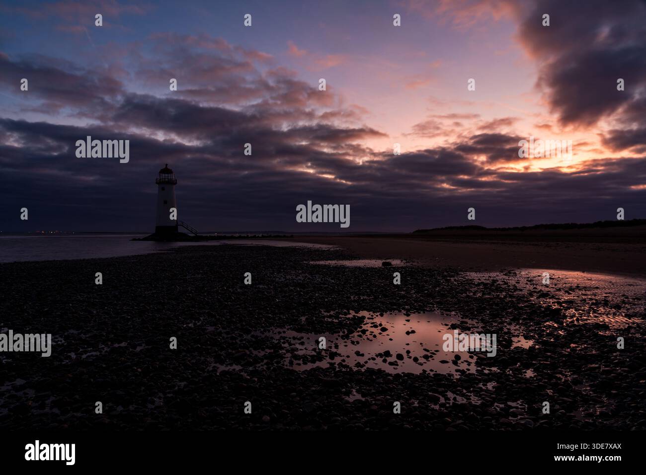 Point of Ayr Lighthouse (Talacre Lighthouse) bei Sonnenaufgang, mit warmem Licht, das über Gezeitenbecken und die Kieselküste unter dramatischer Wolke reflektiert Stockfoto