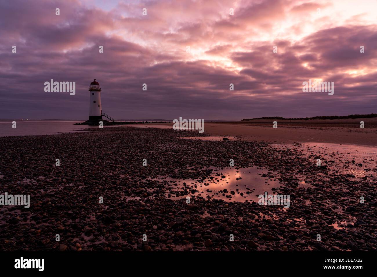 Point of Ayr Lighthouse (Talacre Lighthouse) bei Sonnenaufgang, mit warmem Licht, das über Gezeitenbecken und die Kieselküste unter dramatischer Wolke reflektiert Stockfoto