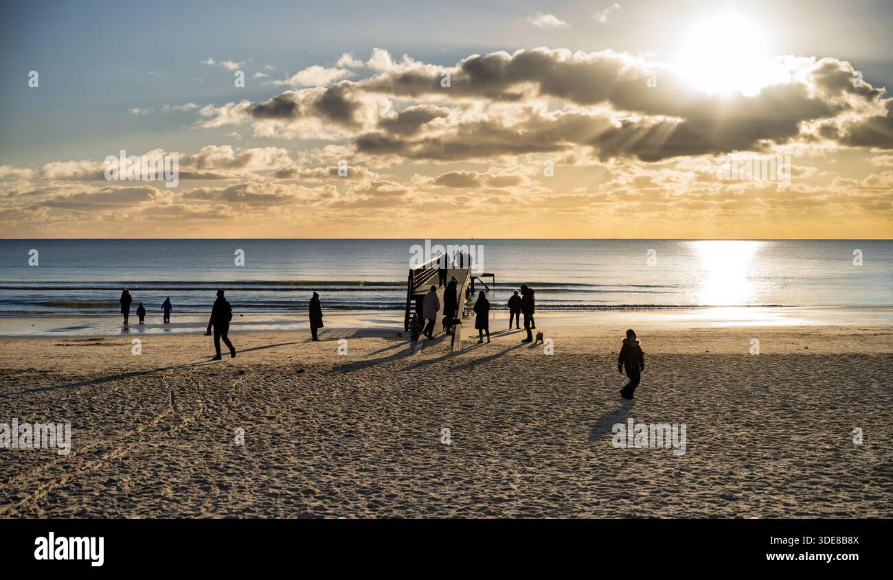 Ein ruhiger Strand bei Sonnenuntergang mit einem hölzernen Pier und Gruppen von Menschen, die entlang der Küste spazieren. Warmes Licht erzeugt lange Schatten und eine ruhige, familiäre fr Stockfoto
