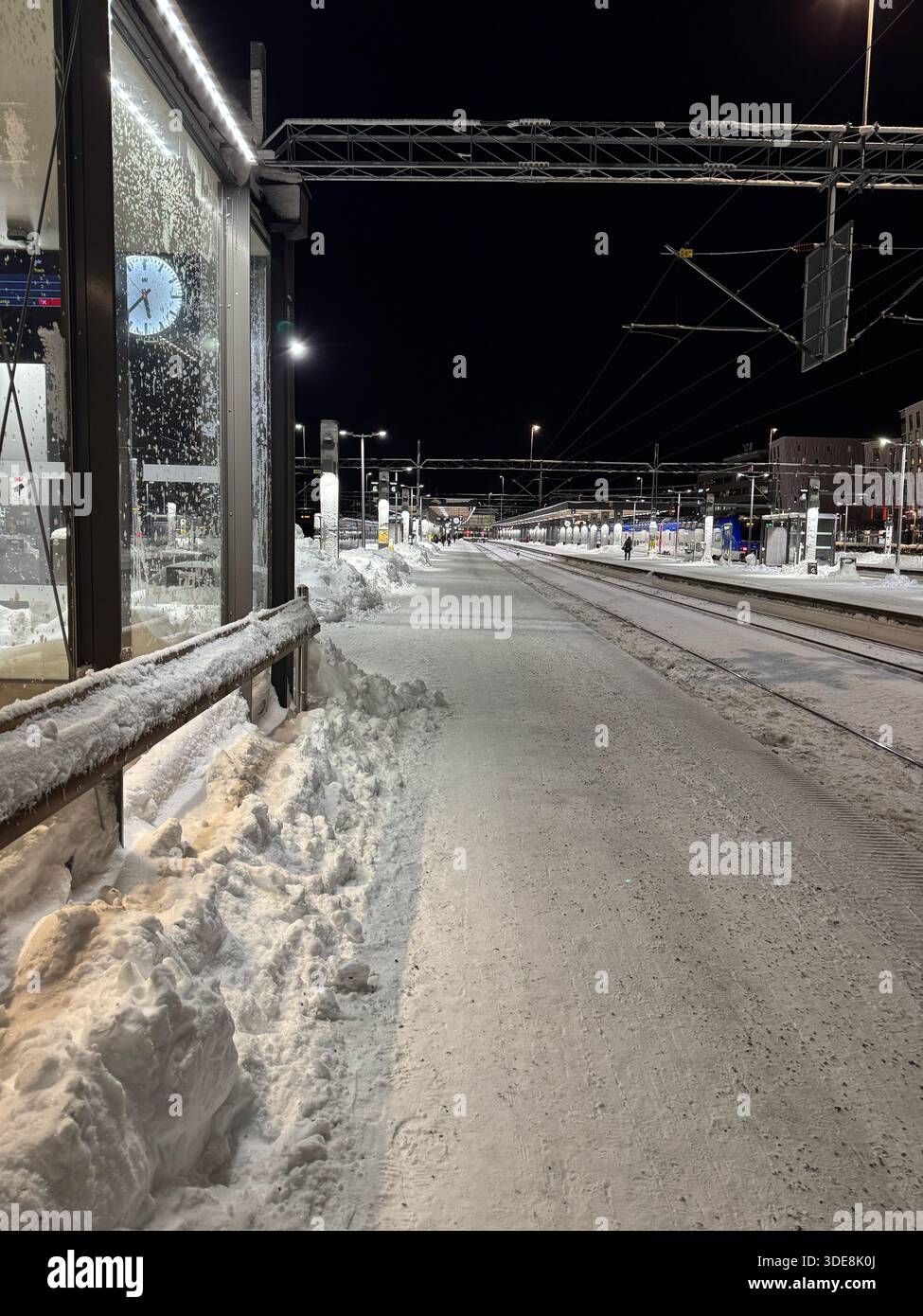 Schneebedeckter Bahnsteig bei Nacht in einer Stadt im Winter, mit leeren Gleisen und beleuchteter Bahnhofsinfrastruktur. - Smartphone-aufgenommenes Stockfoto