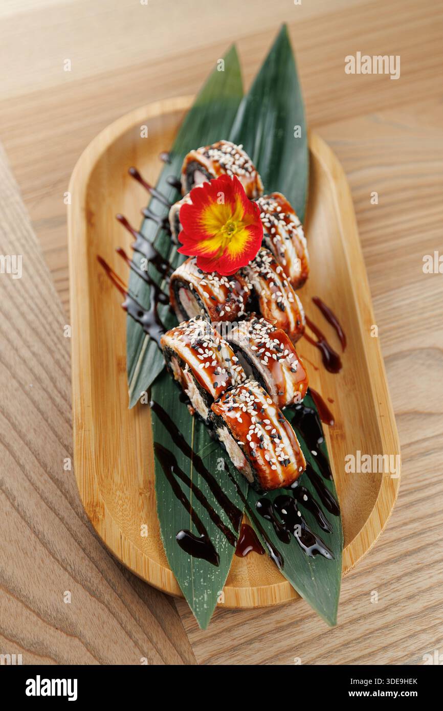 Sushi-Brötchen sind kunstvoll auf einem Bambusteller mit Blättern und einer lebendigen Blume angeordnet. Ein Spritzer Soße sorgt für Kontrast zur Präsentation. Stockfoto