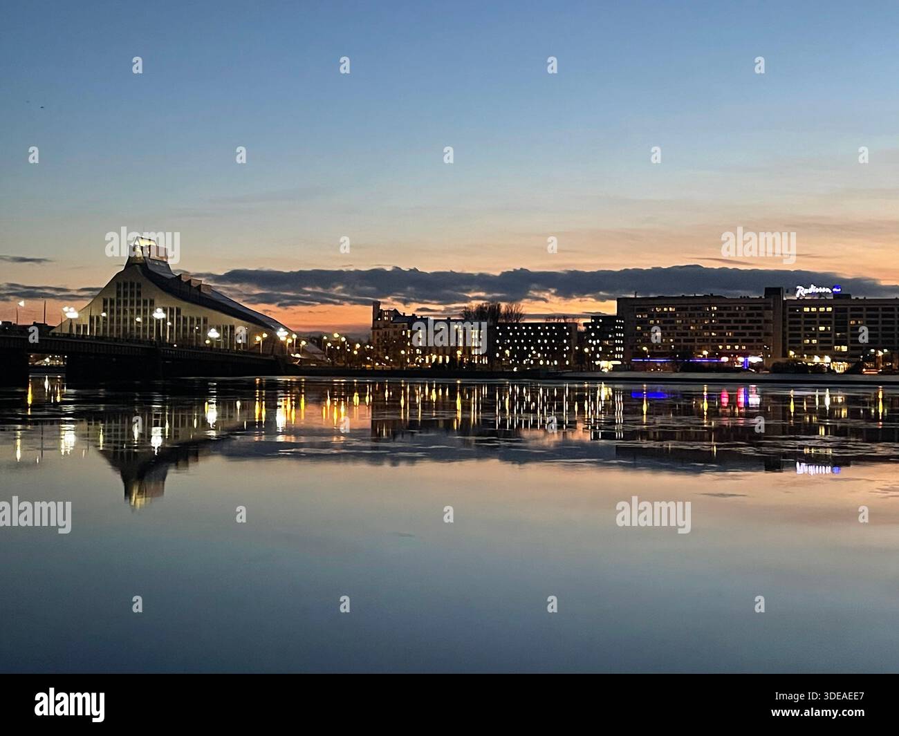Nationalbibliothek Lettlands bei Sonnenuntergang in Riga, mit Stadtlichtern und Reflexionen auf ruhigem Wasser - Smartphone-aufgenommenes Stockfoto
