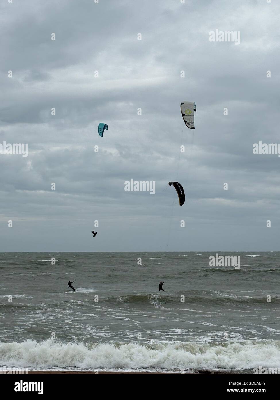 Kitesurfer reiten Wellen unter bewölktem Himmel und machen Sprünge und Manöver in abgehacktem Meer - Smartphone-aufgenommenes Stockfoto