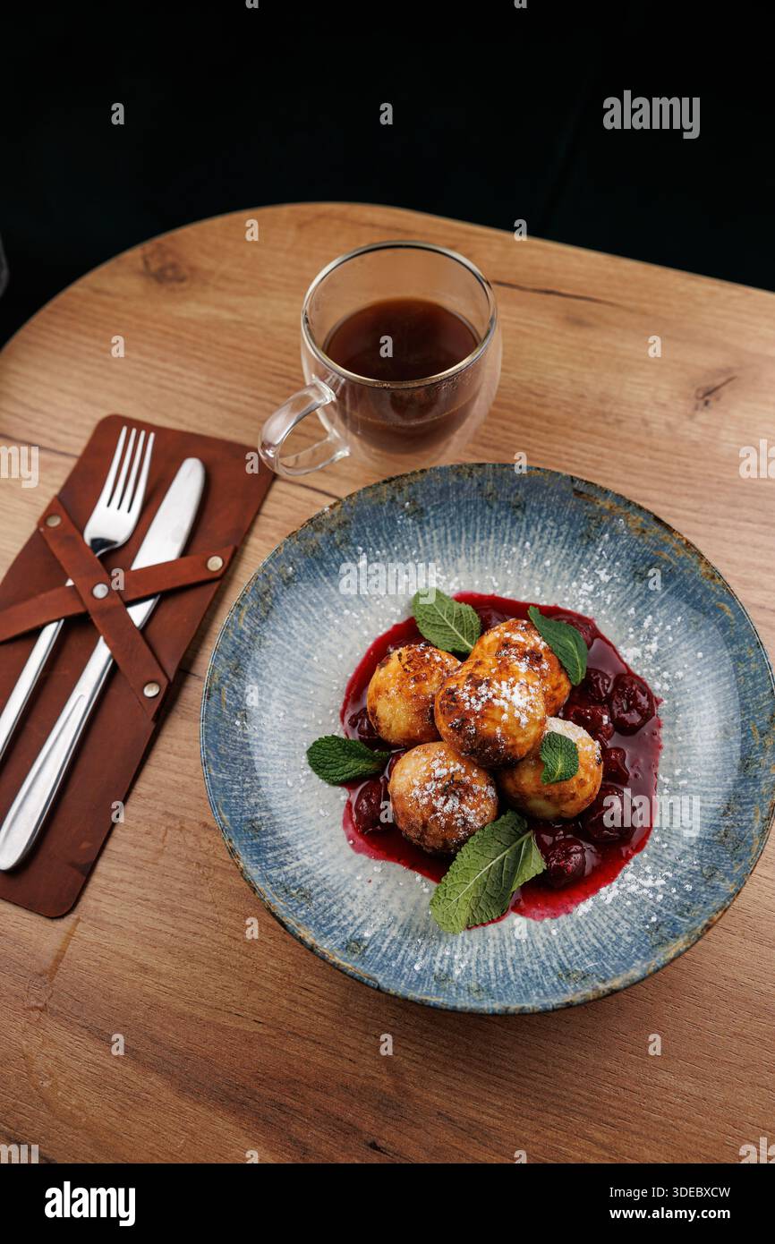 Eine Schüssel mit Süßkäseknödeln in Kirschsauce, garniert mit Minze. In der Nähe liegt eine Tasse Tee auf einem Holztisch mit Besteck. Stockfoto