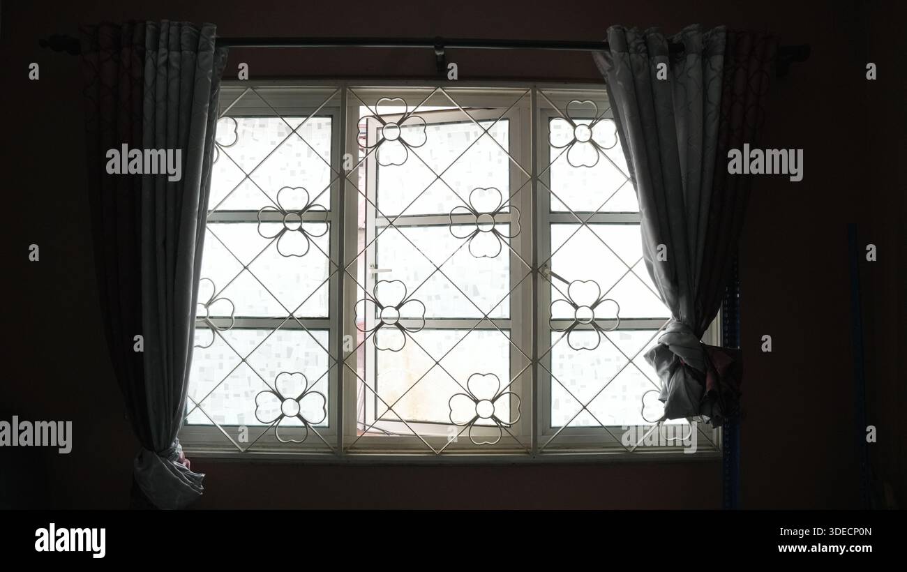 Fenster mit dekorativem Metallgitter und gezogenen Vorhängen lassen Tageslicht in einen dunklen Innenraum einströmen. Stockfoto