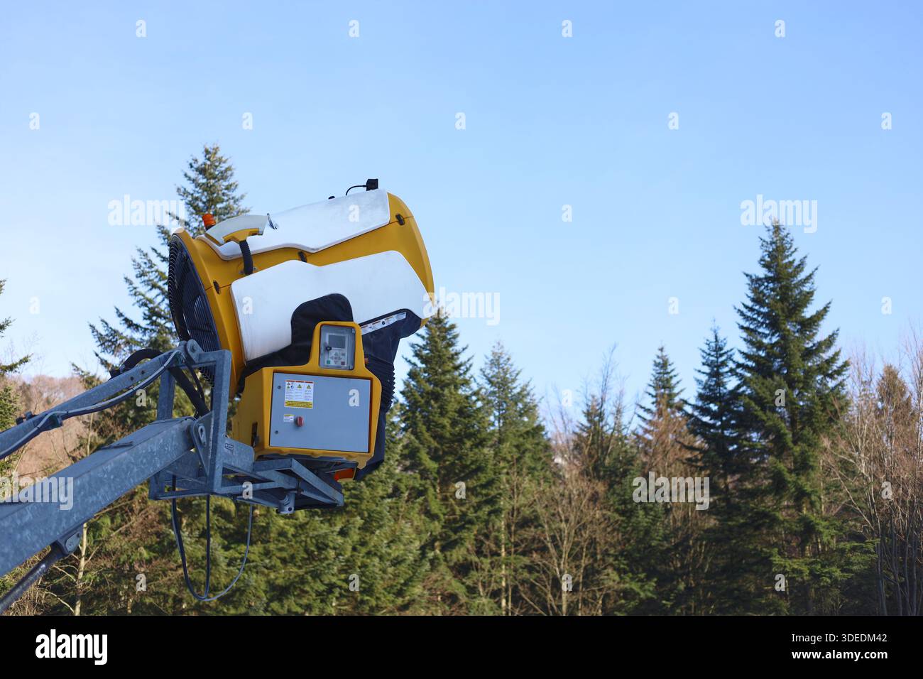 Seitenansicht einer weißen und gelben Schneekanone mit Wald und blauem Himmel Hintergrund Stockfoto