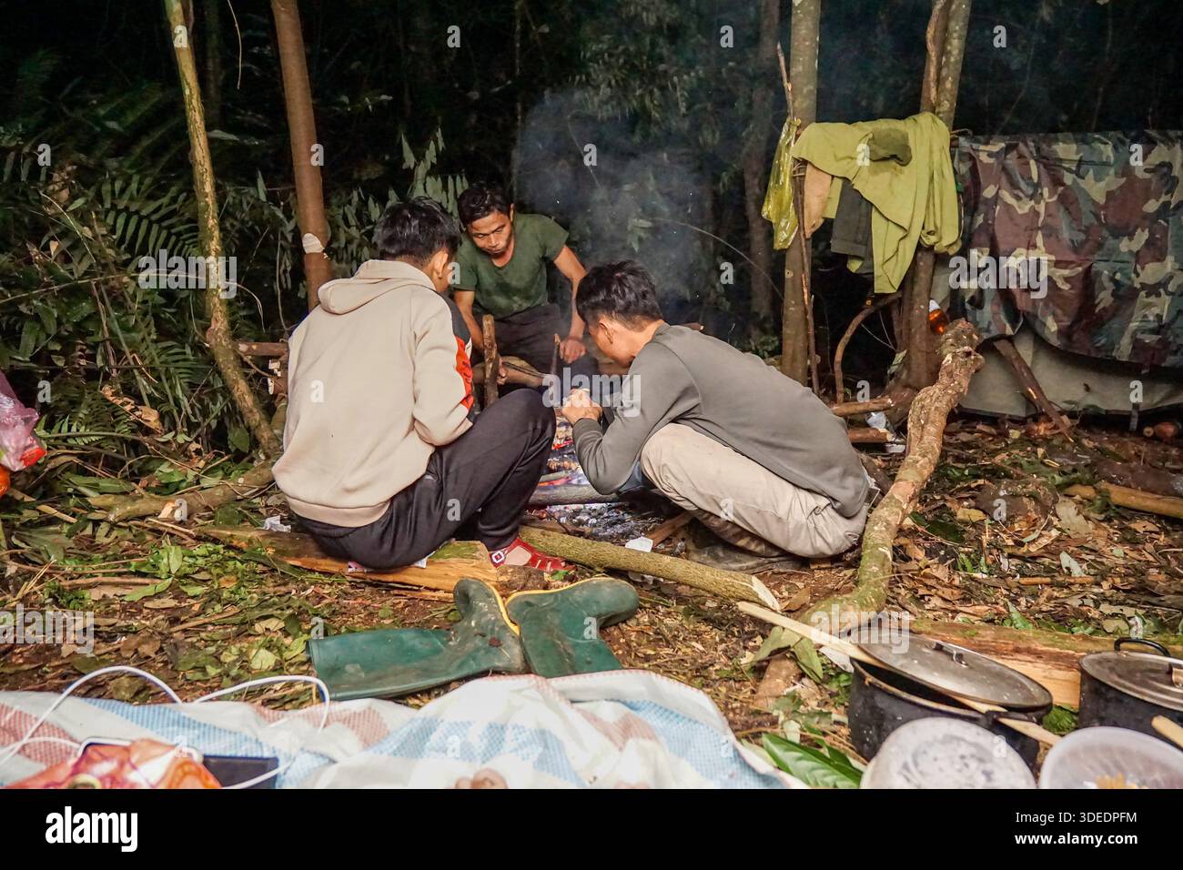 Männer versammeln sich nachts in einem dichten Dschungel um ein knisterndes Lagerfeuer, kochen und teilen Wärme während einer Überlebensexpedition. Stockfoto