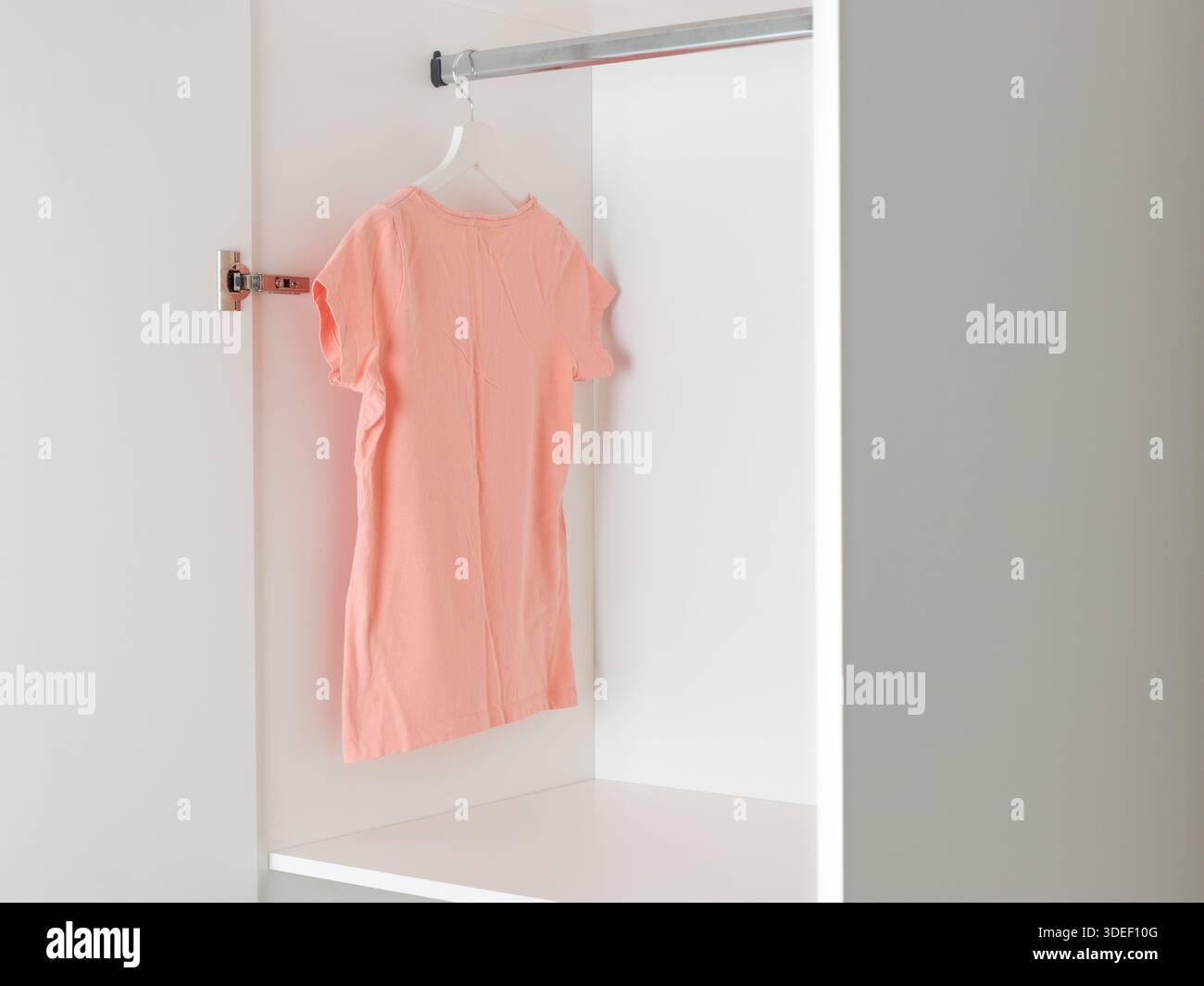Einfarbiges pfirsichfarbenes T-Shirt, das an einem hölzernen Kleiderbügel in einem weißen Kleiderschrank hängt. Minimalistisches Konzept für die Kapselgarderobe, das vielseitige, neutrale und unverzichtbare Elemente hervorhebt Stockfoto