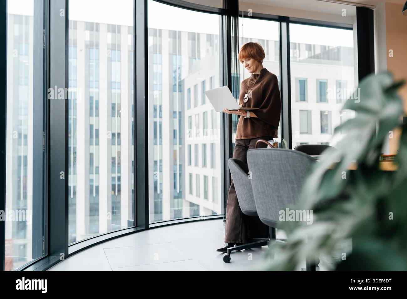 Rundumsicht auf unabhängige Beraterin der 50er-Jahre, die am Laptop in der Nähe des Bürofensters arbeitet. Fokussiert, Vertrauen, digitaler Workflow, modernes Coworki Stockfoto