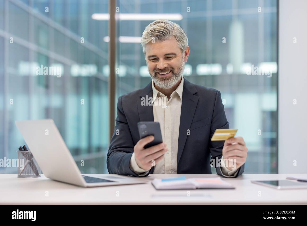 Lächelnder reifer Geschäftsmann am Schreibtisch mit Kreditkarte und Smartphone, während er eine Online-Zahlung oder einen E-Commerce-Kauf durchführt, selbstbewusst und entspannt beim Internet-Banking Stockfoto
