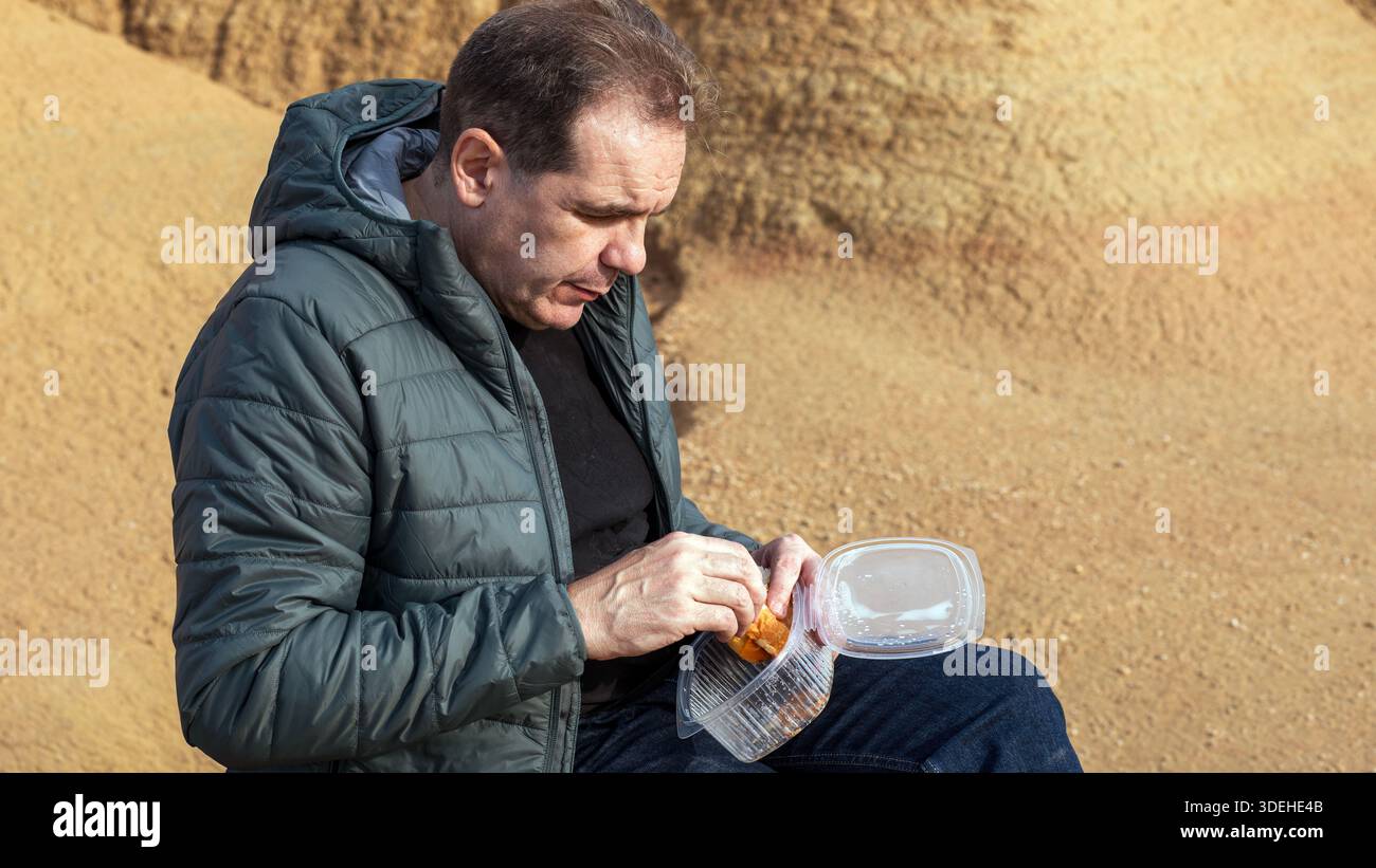 Ein Mann sitzt friedlich auf einem sandigen Hügel in Bardenas Reales. Er packt einen Snack aus, während er von einzigartigen, kargen Landschaften umgeben ist. Die ruhige Atmosphäre ist einzigartig Stockfoto