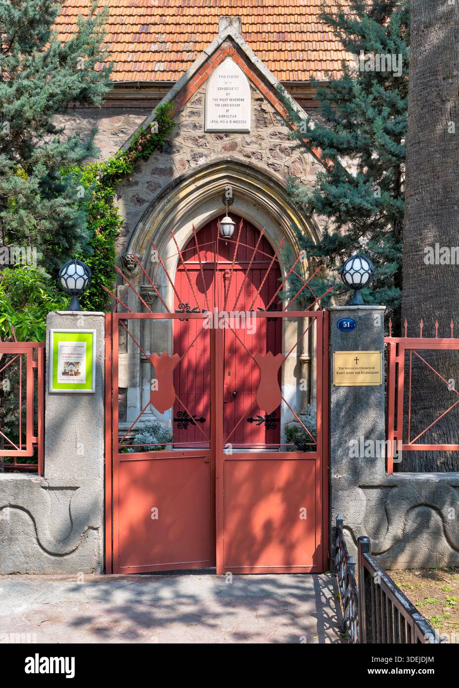Eingang der Anglikanischen Kirche St. Johannes des Evangelisten und der Gedenkhalle des Bischofs Collins, Izmir, Türkei. Mit einer roten Tür, einem kunstvollen Tor, einer Steinfassade, einem gekachelten Dach und grünen Bäumen. Stockfoto