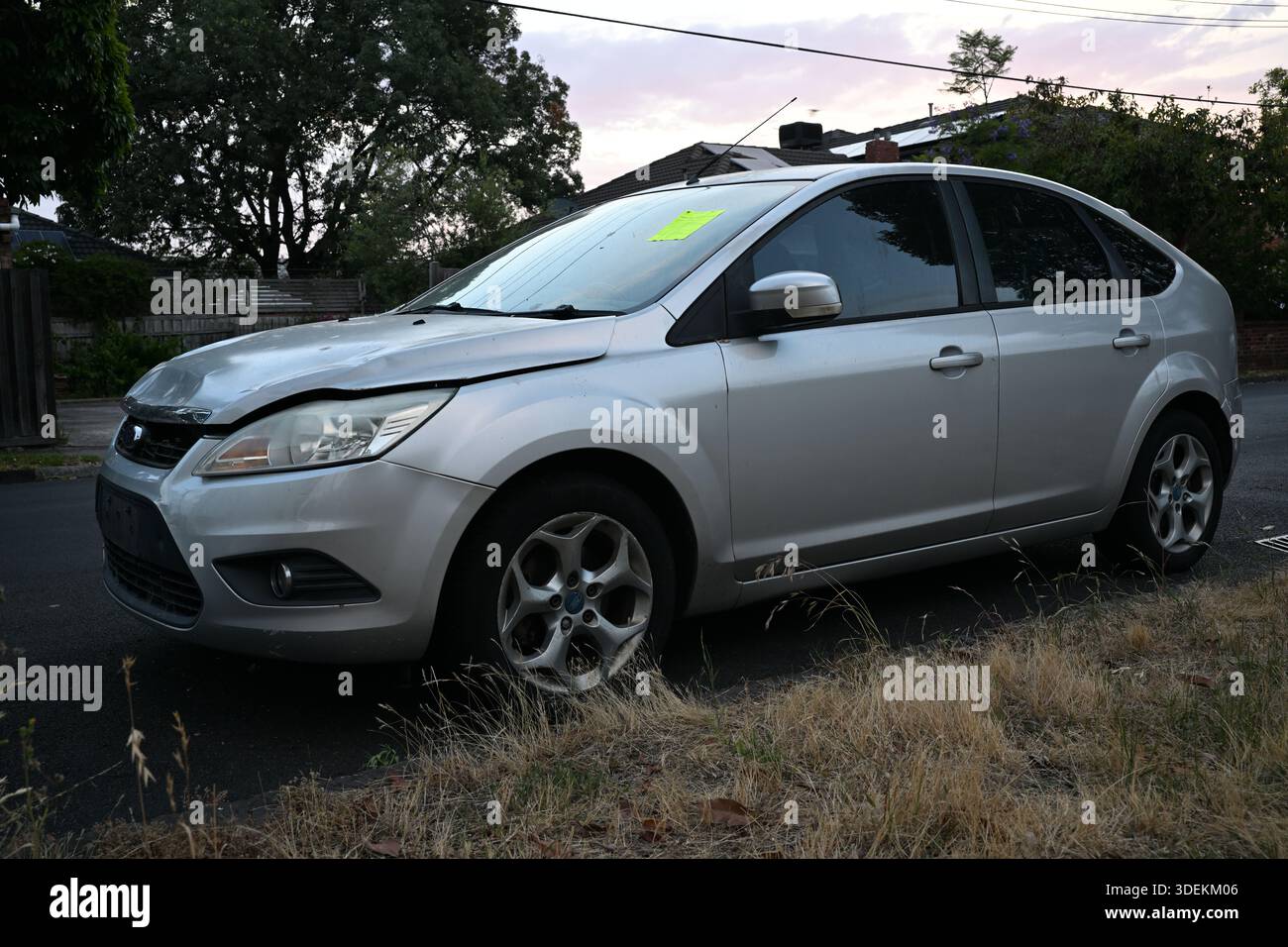 Seitenansicht eines silbernen Ford Focus Autos mit beschädigter Motorhaube und gelbem Schild auf der Windschutzscheibe, das neben langem, trockenem Gras in einer Vorstadtstraße geparkt ist Stockfoto