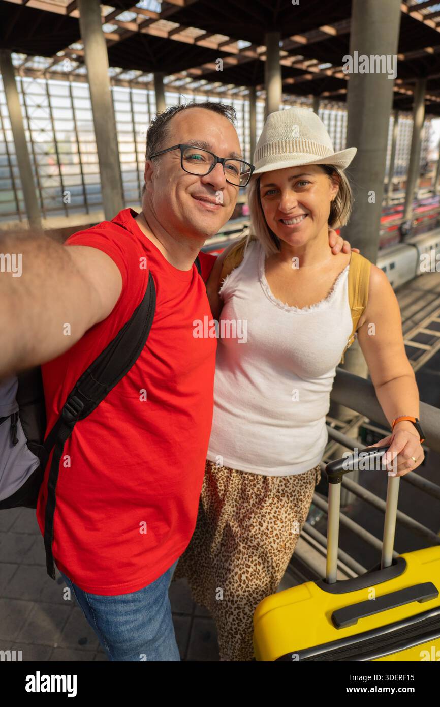 Glückliches, reifes Paar, das mit einem Handy an einem Bahnhof ein Reisegedächtnis aufnimmt. Sie reisen zusammen mit Gepäck und Reisedokumenten. Stockfoto