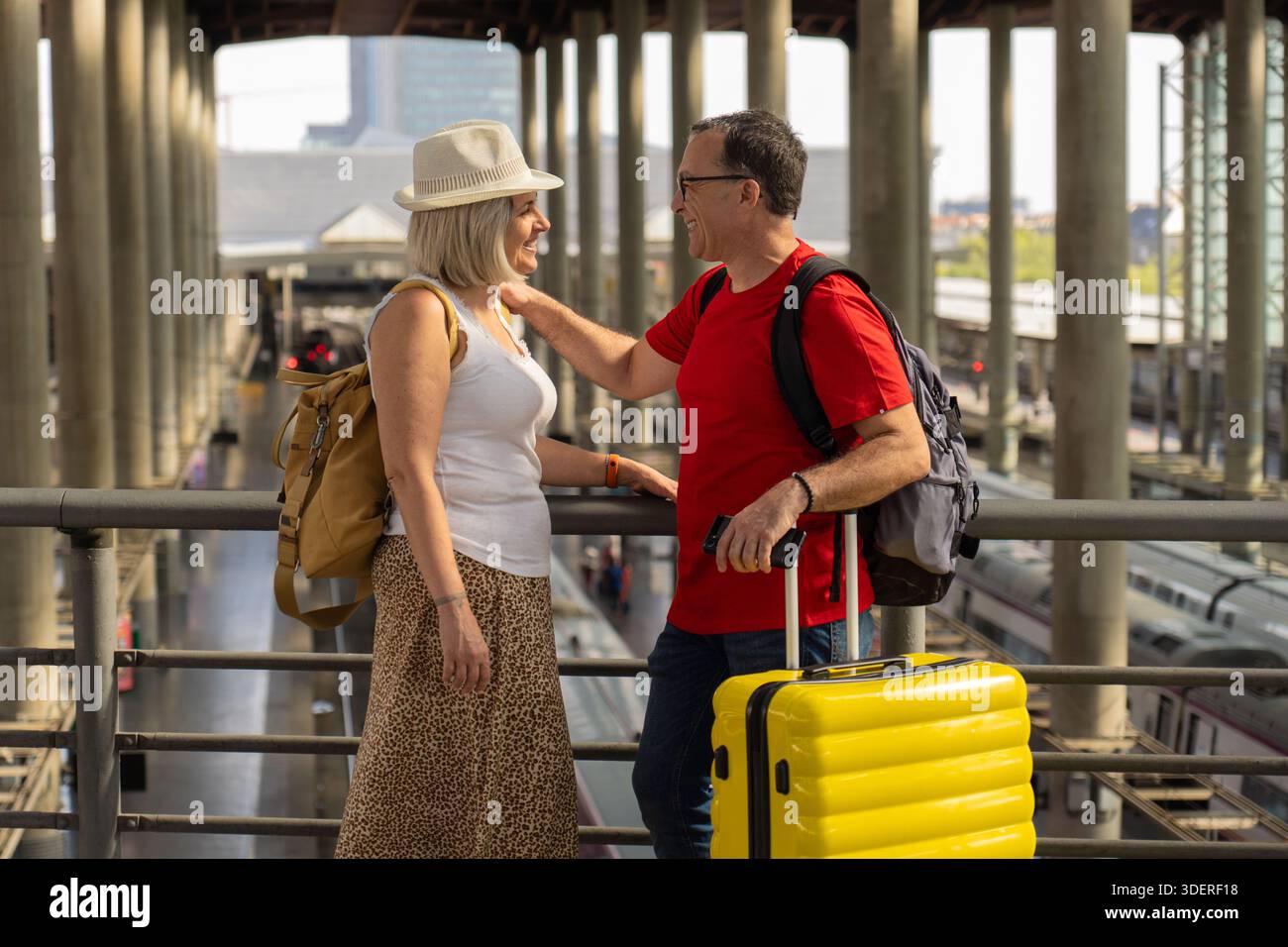 Glückliches reifes Touristenpaar, das in einem großen Bahnhof mit moderner Architektur und den Zügen darunter steht. Sie lächeln und genießen ihre Freude Stockfoto
