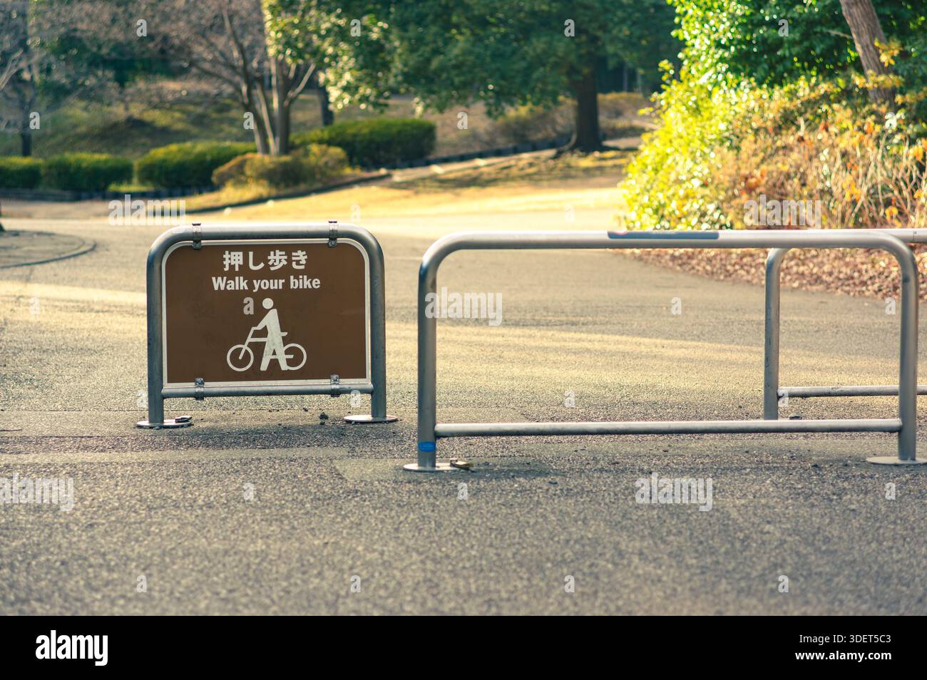 Ein japanisches und englisches Schild weist die Besucher an, mit dem Fahrrad in einem Park zu spazieren. Der Weg ist klar mit Bäumen und Grünflächen, die ihn umgeben. Stockfoto