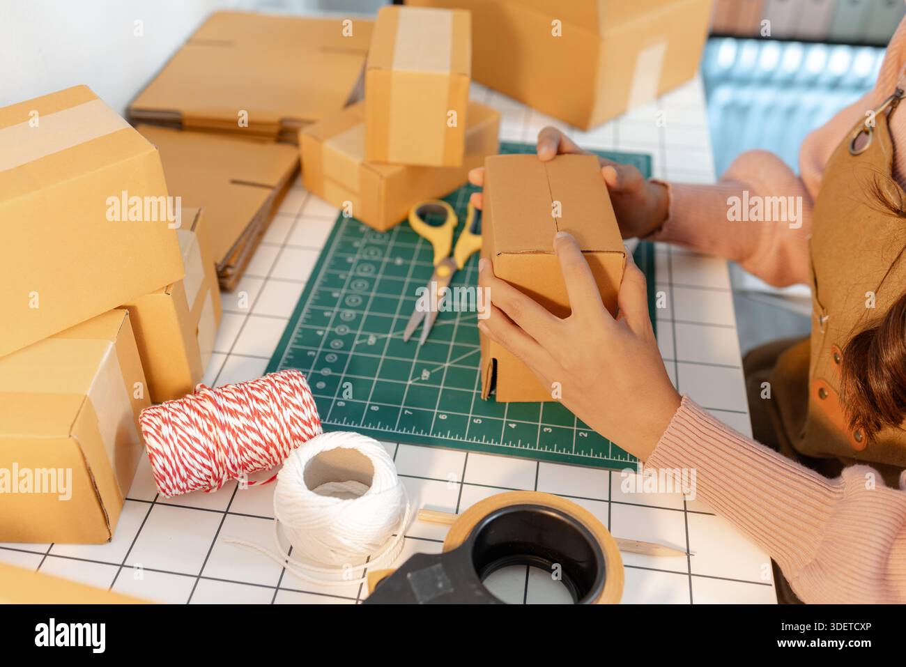 Eine junge Frau baut sorgfältig Pappkartons an einem gut organisierten Arbeitsplatz zusammen. Der Schreibtisch ist mit Verpackungsmaterial, Klebeband und Schnur gefüllt Stockfoto