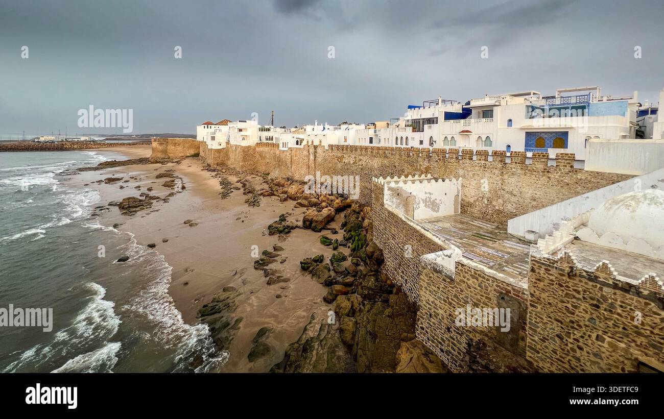 An einem regnerischen Tag krachen Meereswellen am Strand am Fuße der Festungsmauern der befestigten Stadt Asilah Stockfoto