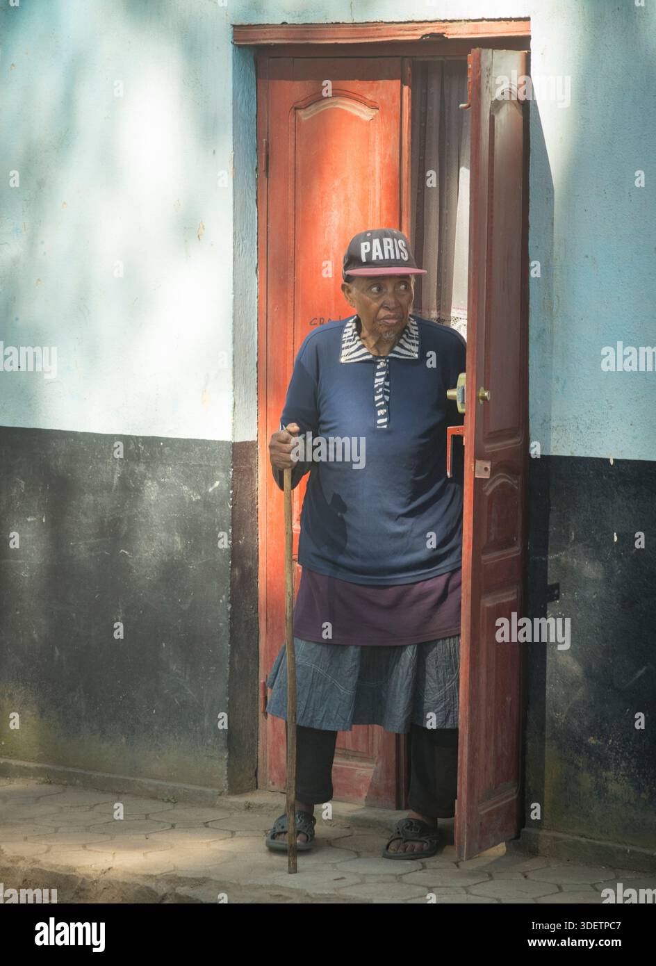 Ein Porträt eines älteren madagassischen Dorfes mit einem Gehstock und einer Baseballmütze, der an der Tür ihres städtischen Hauses in Ambatolampy, Madagaskar, steht. Stockfoto