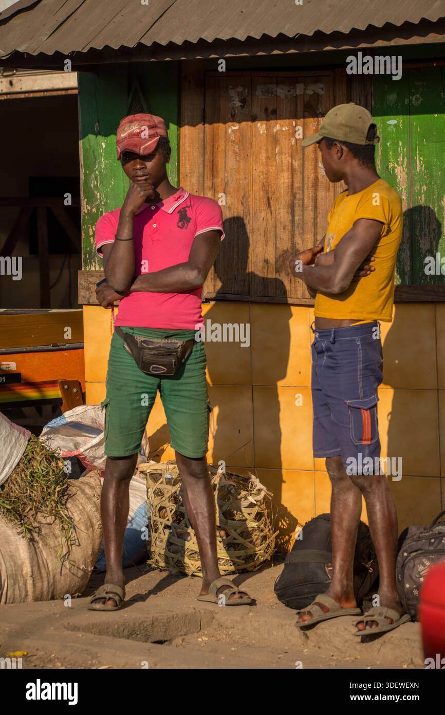 Die Szene junger madagassischer Männer in farbenfrohen, modernen Kleidern und Baseballmützen auf dem Dorfmarkt von Alakamisy Ambohimaha veranschaulicht die Verschmelzung globaler Modetrends mit traditioneller madagassischer Kultur und dem lebhaften Gemeinschaftsleben in den zentralen Hochländern. Die Einführung zeitgenössischer Kleidung spiegelt den Einfluss internationaler Trends wider, die selbst abgelegene Dörfer über die Märkte für Gebrauchtkleidung und die Medienpräsenz erreichen. Im Gegensatz zu traditionelleren lamba-Tüchern ist es eine Möglichkeit für junge Menschen, ihre moderne Identität auszudrücken und gleichzeitig am Gemeinschaftsleben teilzunehmen Stockfoto