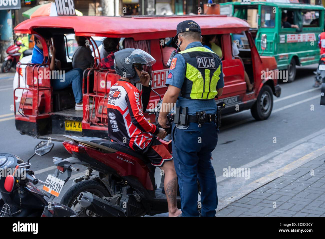 Der Polizist führt eine Straßenkontrolle eines Motorradfahrers durch, während Fahrzeuge mit öffentlichen Verkehrsmitteln eine geschäftige Straße entlang fahren. Stockfoto