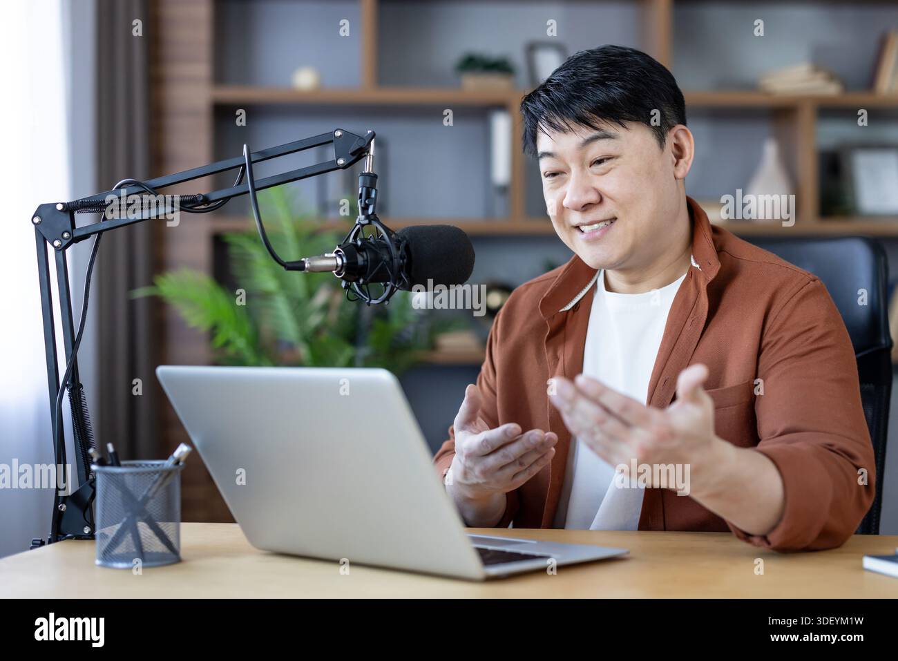 Asiatischer Mann, der ansprechende Inhalte in seinem Heimstudio erstellt, in ein professionelles Mikrofon spricht und einen Laptop ansieht, während er sendet oder rekordiert Stockfoto
