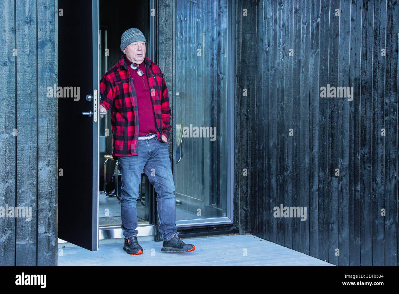 Ein älterer Mann in kariertem Hemd und Beanie steht vor der Tür und unterstreicht die moderne Holzarchitektur und das einladende Ambiente des Heims, betont Wärme und Stockfoto