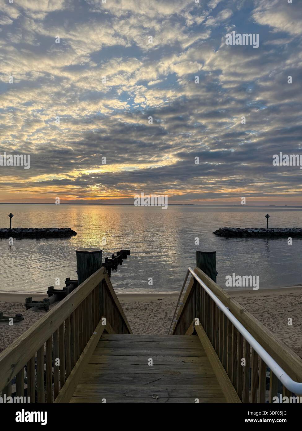 Eine hölzerne Strandtreppe führt bei Sonnenaufgang zu einer ruhigen Bucht mit goldenen Reflexen auf dem Wasser unter dramatischen Morgenwolken. - Smartphone-aufgenommenes Stockfoto