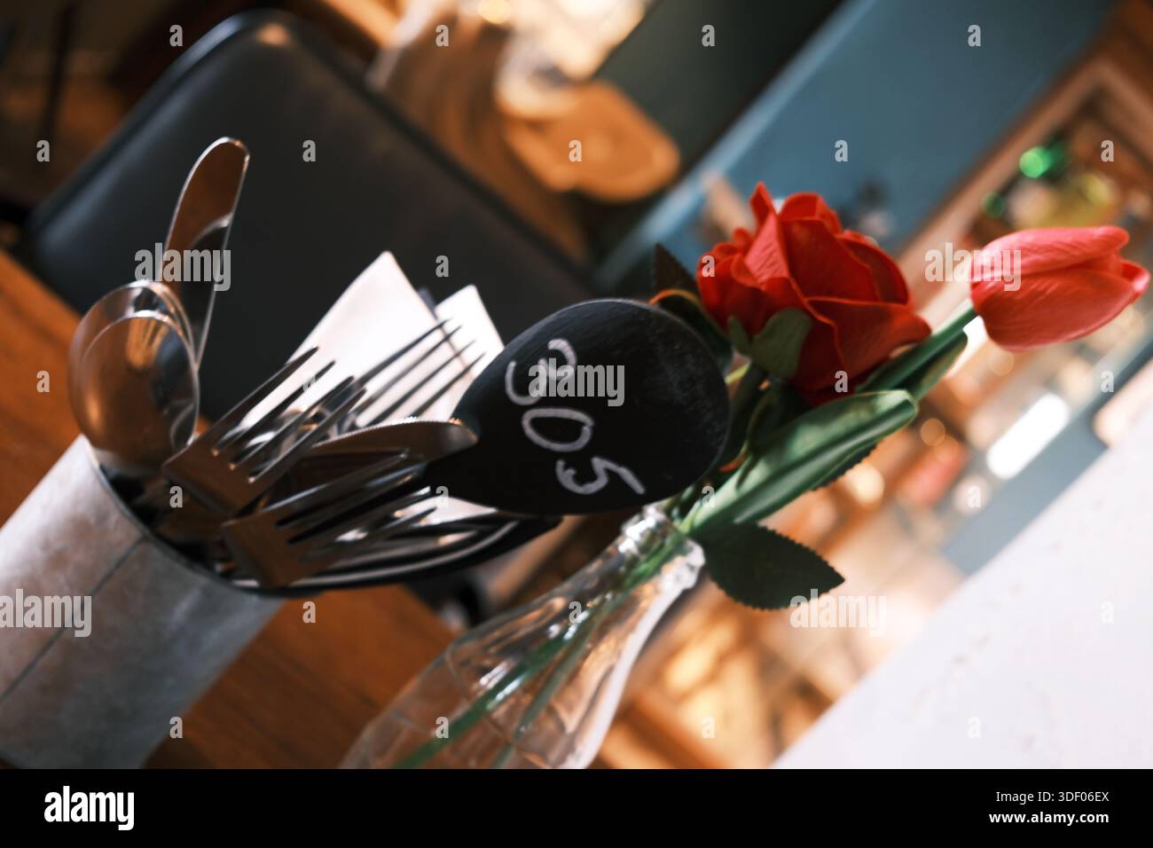 Gehobener Bistro-Tisch mit Besteck, roter Rose und Tulpe in einer Glasflasche, geringe Tiefe des Feldes, warmes Restaurant-Ambiente im Inneren. Stockfoto