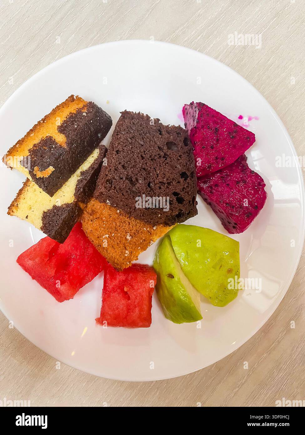Verschiedene Scheiben Kuchen und frische tropische Früchte einschließlich Wassermelone, Drachenfrucht und Guave auf einem weißen Teller malaysia - Smartphone-aufgenommenes Stockfoto