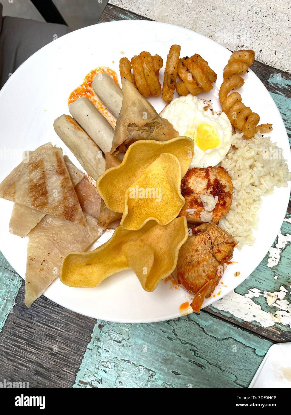 Teller mit verschiedenen asiatischen und westlichen Speisen wie Reis, Spiegelei, Würstchen, Huhn, Samosas, und Chips auf rustikalem Tisch malaysia - Smartphone-aufgenommenes Stockfoto