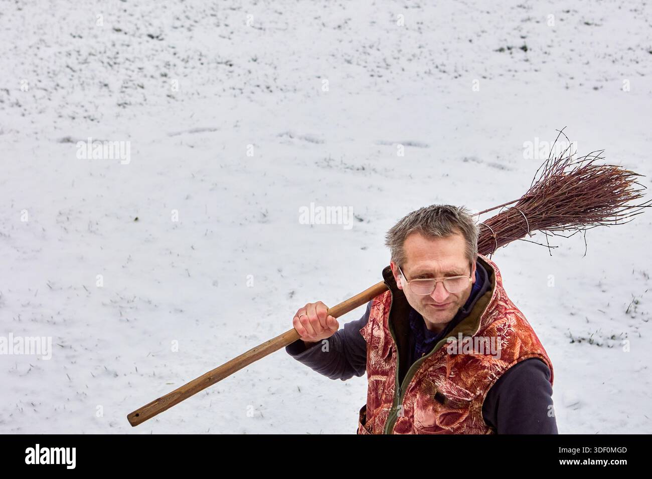 Der Oberkörper zeigt eine Erwachsene Figur mit Holzbesen auf der Schulter vor verschneiten Hintergrund mit weggerichtetem Blick und einer komponierten Haltung. Stockfoto