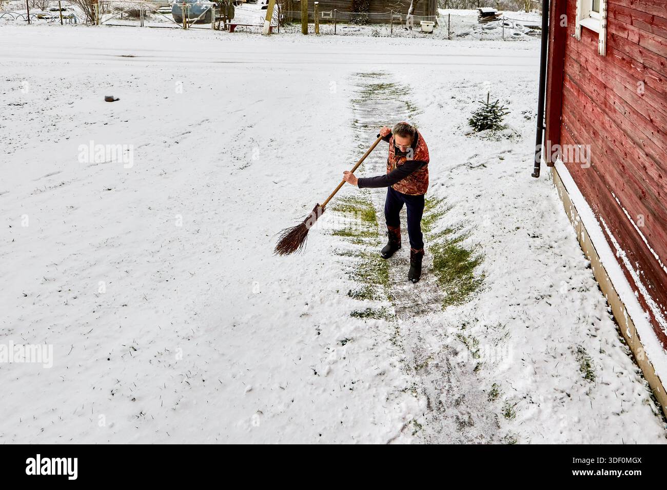 Der Mann räumt Schnee vom Betonsteg zum Eingang des Landhauses mit einem Besen, der eine isolierte Weste und Pelzstiefel trägt. Stockfoto