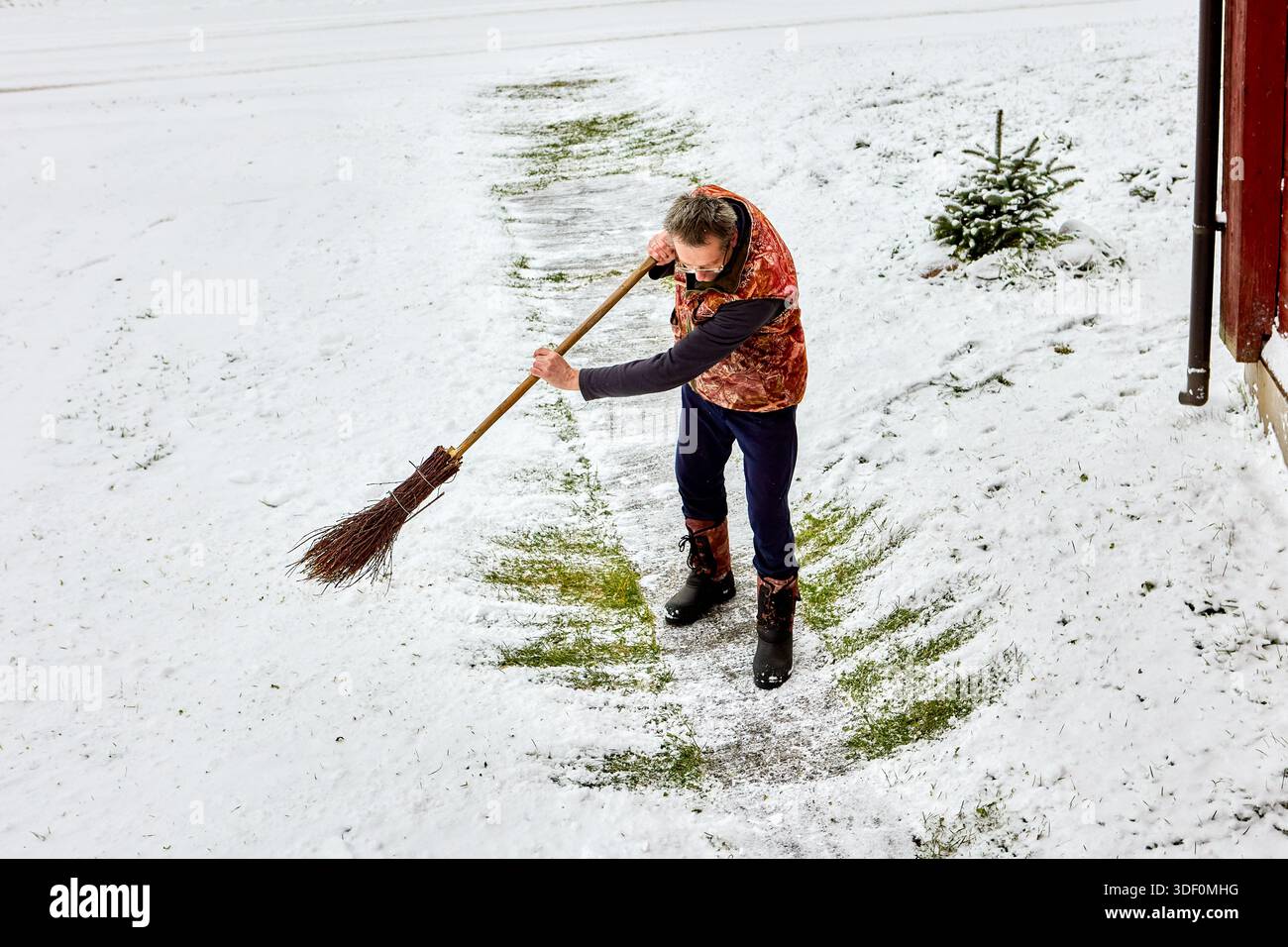 Verwendung von Besen zum Kehren von Schnee vom Betonsteg zur Eingangstür des ländlichen Hauses Mittelalter Mann in leichter Winterkleidung fegt Hof. Stockfoto