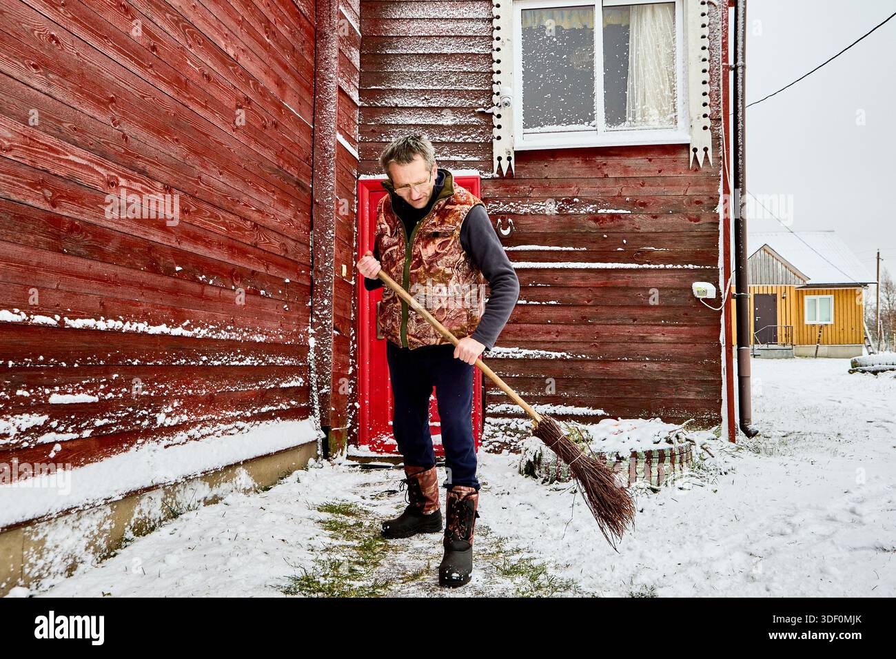 Der reife Mann fegt Schnee vom Eingangsweg mit Birkenbesen und führt tägliche Winterarbeiten durch, um den Bereich vor dem Landhaus mit lackiertem Re zu pflegen Stockfoto