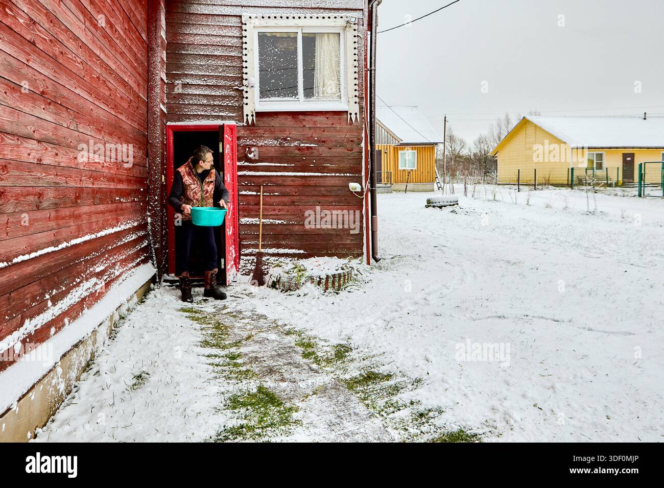 Reifer Mann verlässt die Tür des Landhauses mit Plastikbecken in den Händen, um nach der Nassreinigung Wasser auf den Schnee im Hof zu gießen. Stockfoto
