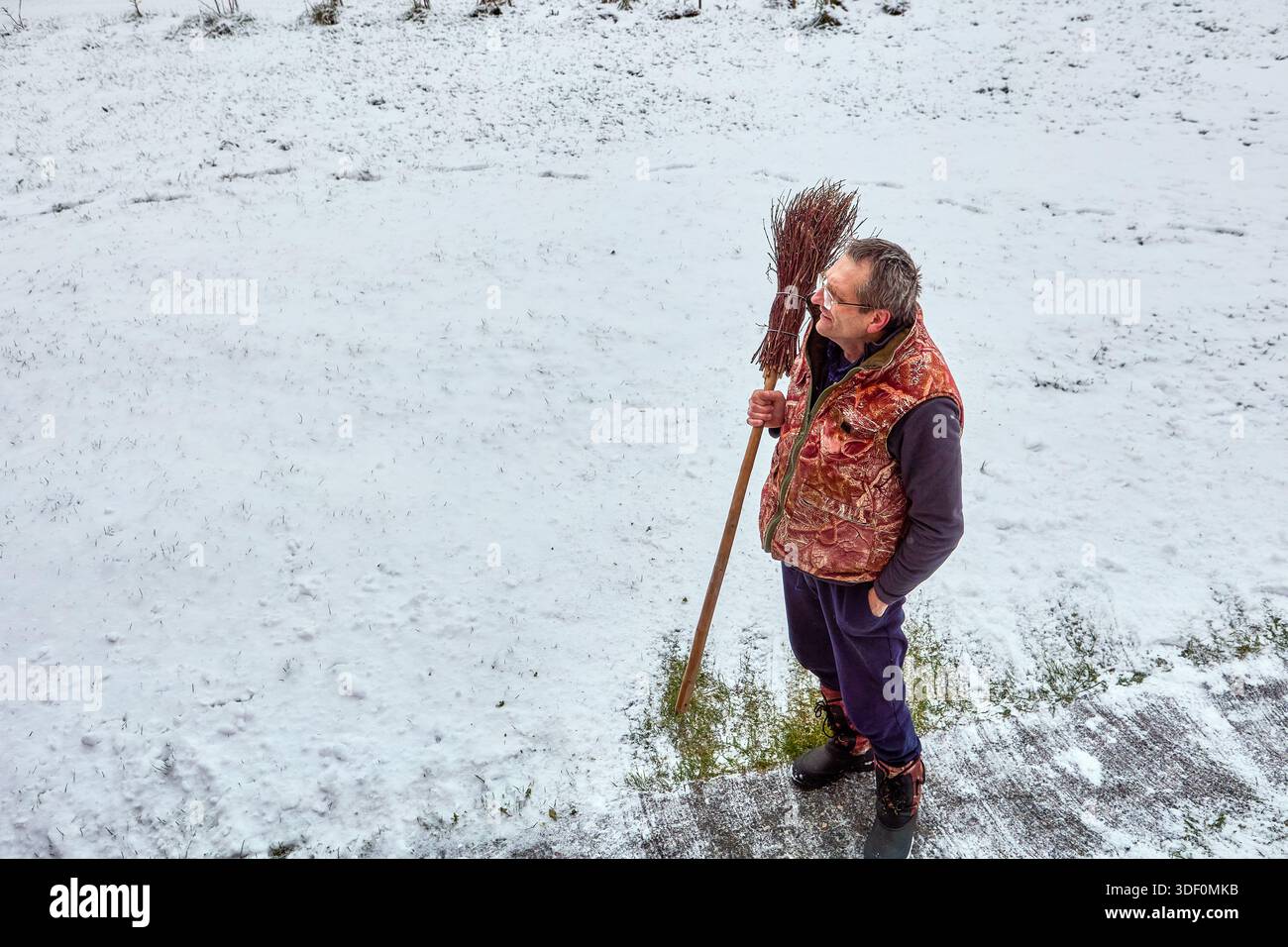 Einsame menschliche Figur hält auf schneebedeckter Oberfläche inne und greift rustikalen Besen in warmer, mehrschichtiger Kleidung, die aus erhöhter Perspektive aufgenommen wurde. Stockfoto