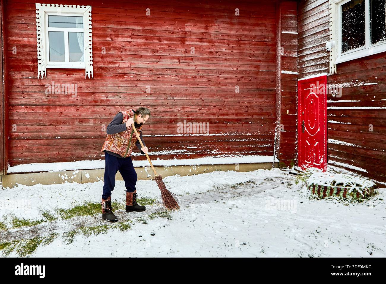 Vor dem mit roten Brettern bekleideten Landhaus fegt reifer Mann mit Birkenbesen den Gartenweg und entfernt Schnee nach Schneefall. Stockfoto