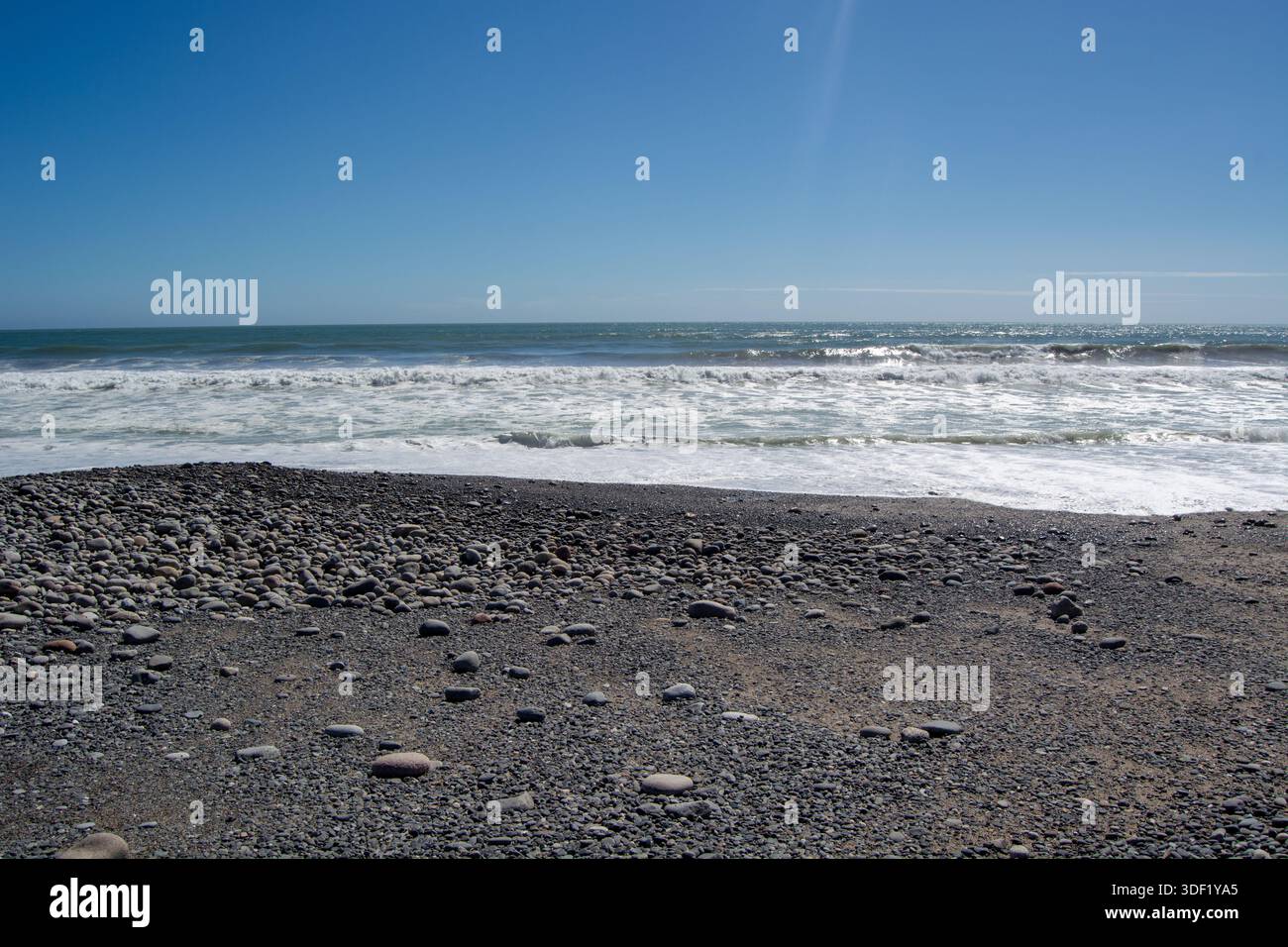 Eine ruhige Strandszene mit einem felsigen Ufer mit glatten Kieselsteinen, sanften Wellen am Wasserrand und einem klaren blauen Himmel über dem Wasser. Stockfoto