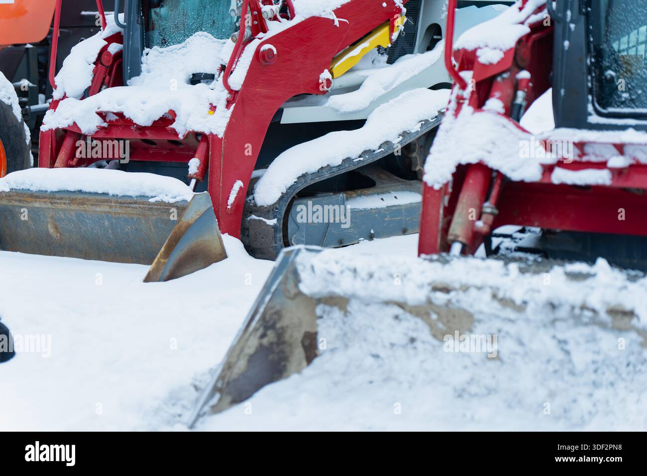 Rote Bauladerschaufeln und Hydraulikarme, die mit Schnee bedeckt sind und auf dem Boden der Winterbaustelle geparkt sind und schwere Maschinen bei kaltem Wetter zeigen Stockfoto