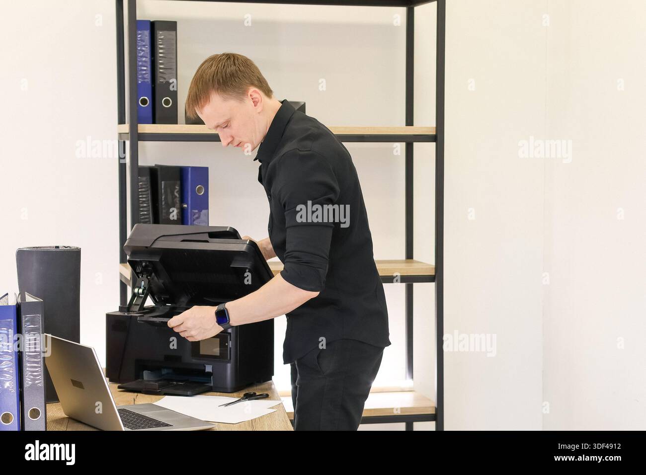 Ein junger Mann in formeller Kleidung greift in den geöffneten Drucker, um Teile in einem ordentlichen Büro mit Regalen zu reparieren oder anzupassen. Stockfoto