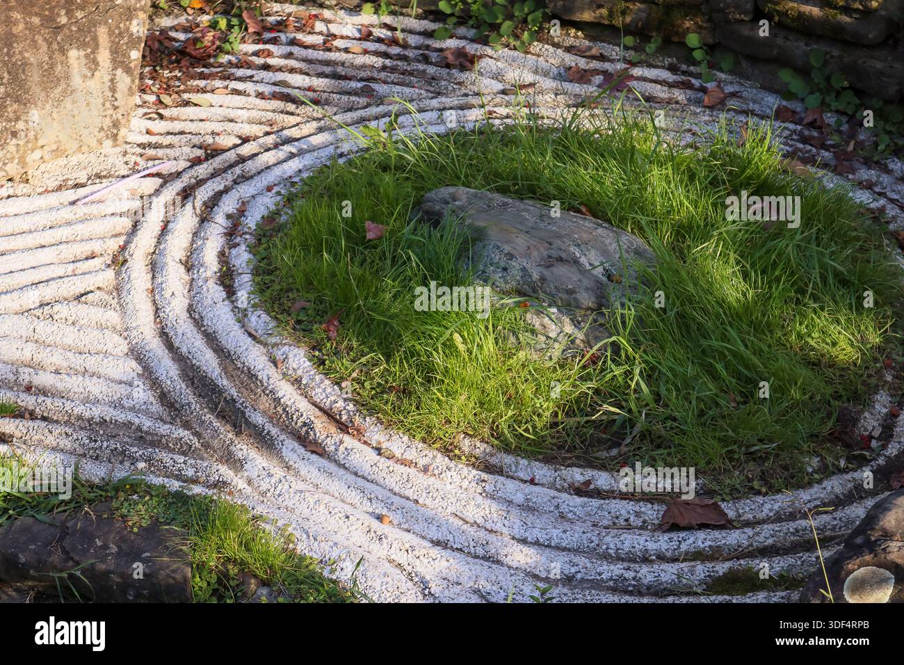 Kompliziertes Zen-Gartendesign mit schwirrenden, weißen Kieswellen um einen zentralen, moosigen Felsbrocken, durchsetzt von leuchtend grünen Grasbüscheln und einer grünen Krautkugel Stockfoto