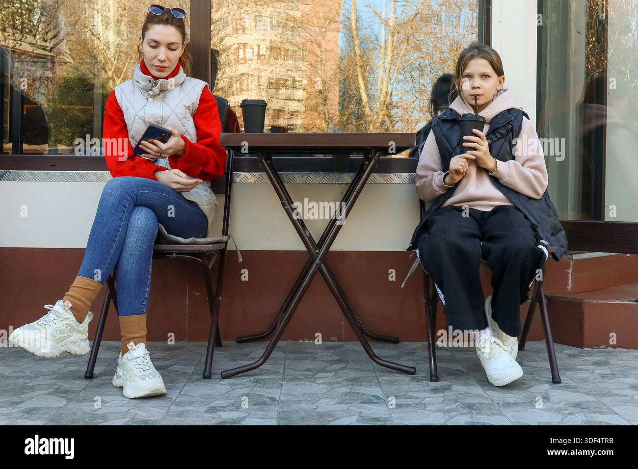 Eine Erwachsene Frau trinkt aus dem Becher und schaut das Telefon, sitzt neben einem Mädchen aus der Jugend und schlürft ein Getränk im Café im Freien in Freizeitkleidung. Stockfoto