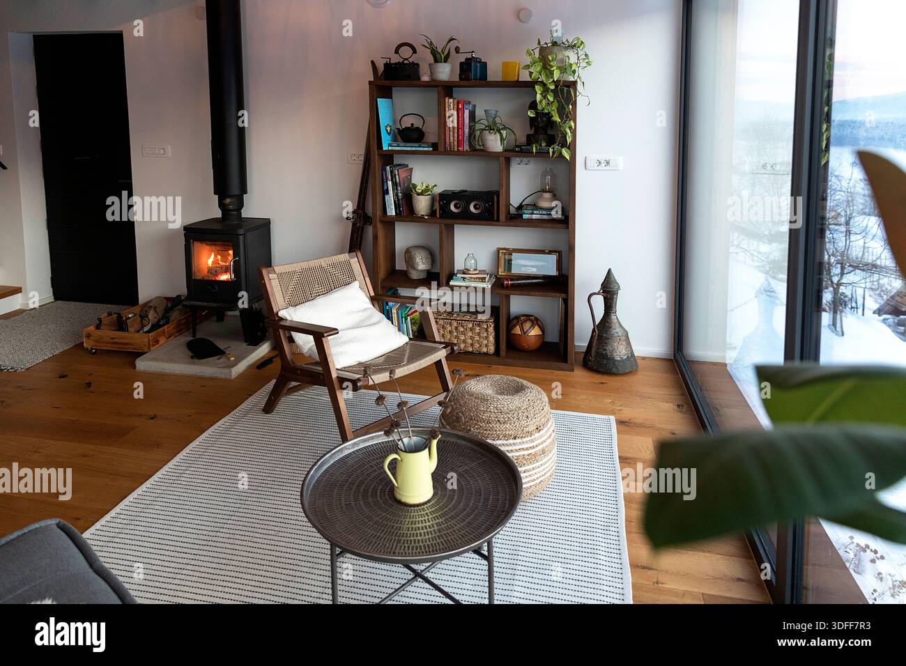 Gemütliches Wohnzimmer im skandinavischen Stil mit Kamin und Panoramafenster mit minimalistischen Möbeln, Pflanzen und malerischem Blick auf die Landschaft Stockfoto