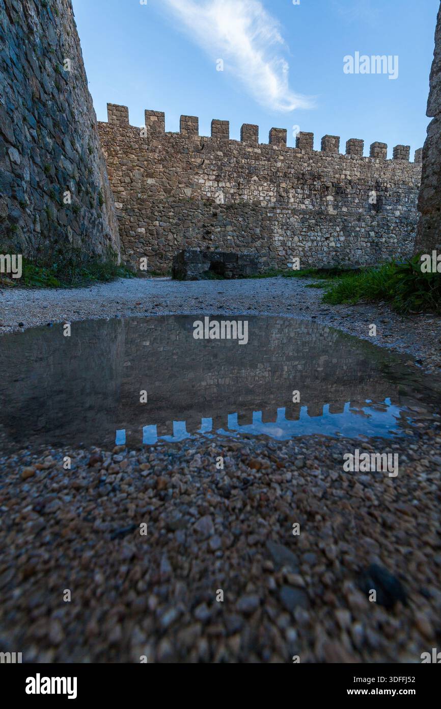 Leiria. Ein Niedrigwinkelfoto fängt die alten Steinmauern einer Burg mit ihren zinnenbesetzten Zinnen ein, die sich perfekt in einer Pfütze auf dem Kies spiegeln Stockfoto