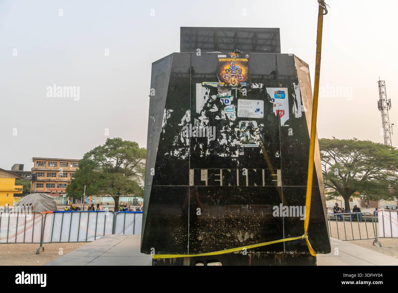 Das Lenin-Denkmal in Cotonou, Benin, ist mit Plakaten und Emblemen umzäunt und erinnert an die kommunistische Geschichte inmitten von Palmen und urbaner Kulisse. Stockfoto